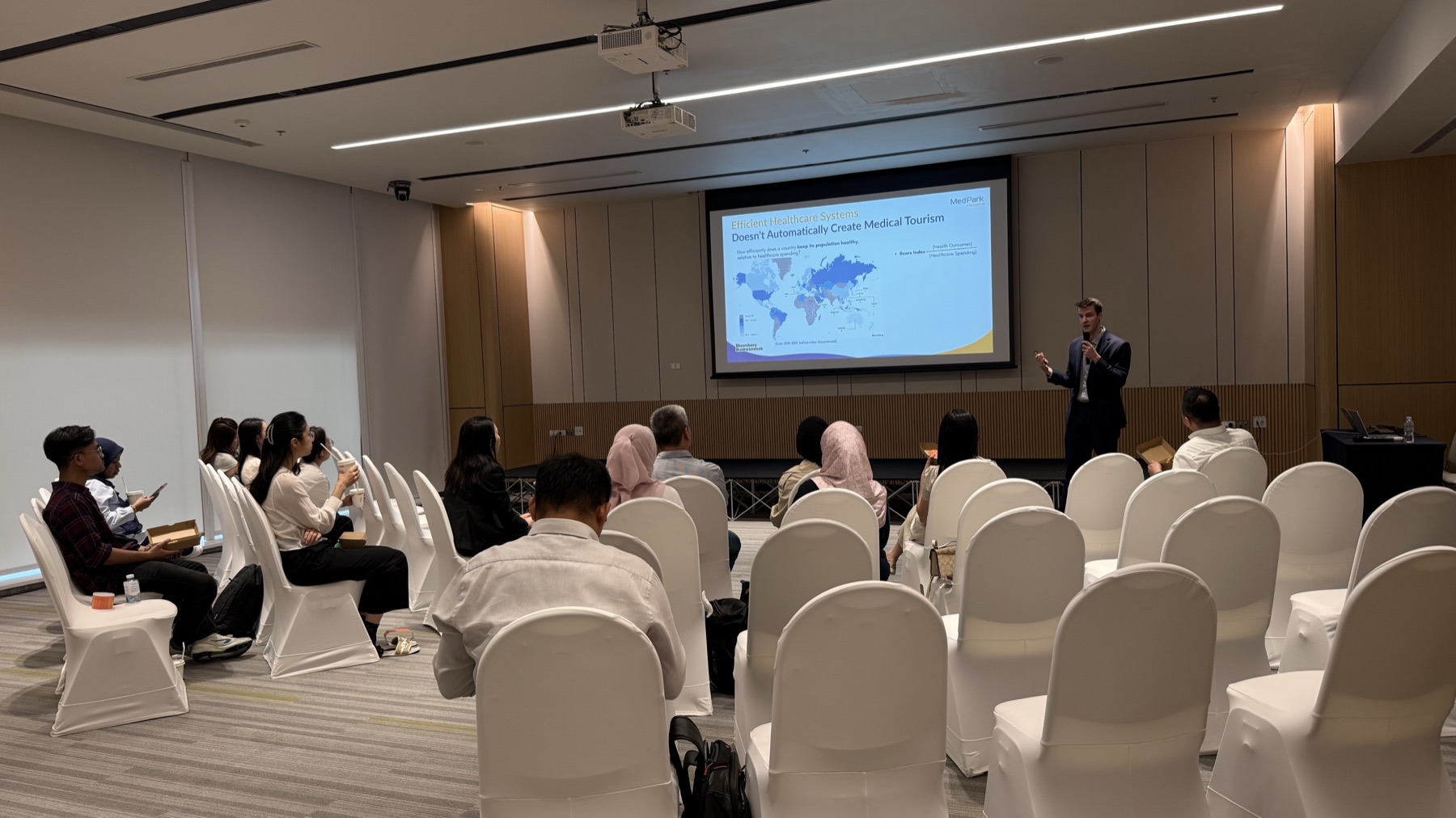 A speaker presenting healthcare-systems data citing Bloomberg Businessweek to a seated audience