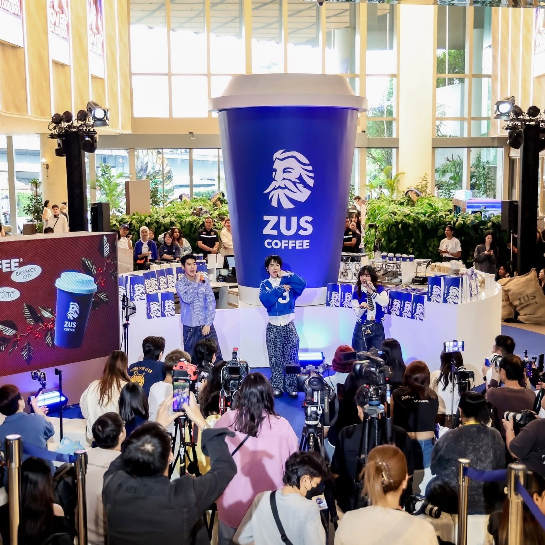 A giant branded coffee cup backdrop and three performers on stage at a ZUS Coffee retail launch event, press and crowd in front